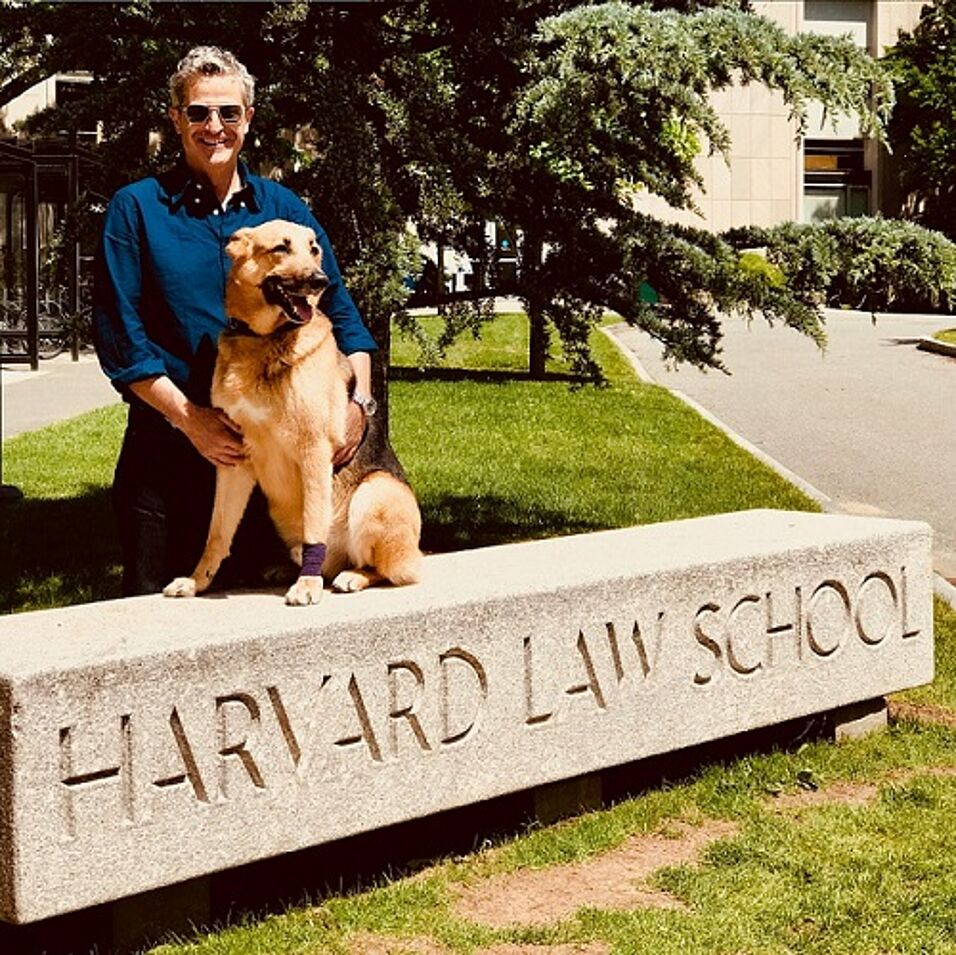 Bild: Foto von Prof. Ebrahim Afsah mit seinem Hund hinter einem Steinschild der Harvard Law School