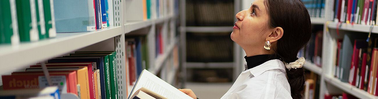 Bild: Eine Studierende mit einem aufgeschlagenen Buch in der Hand in einer Bibliothek