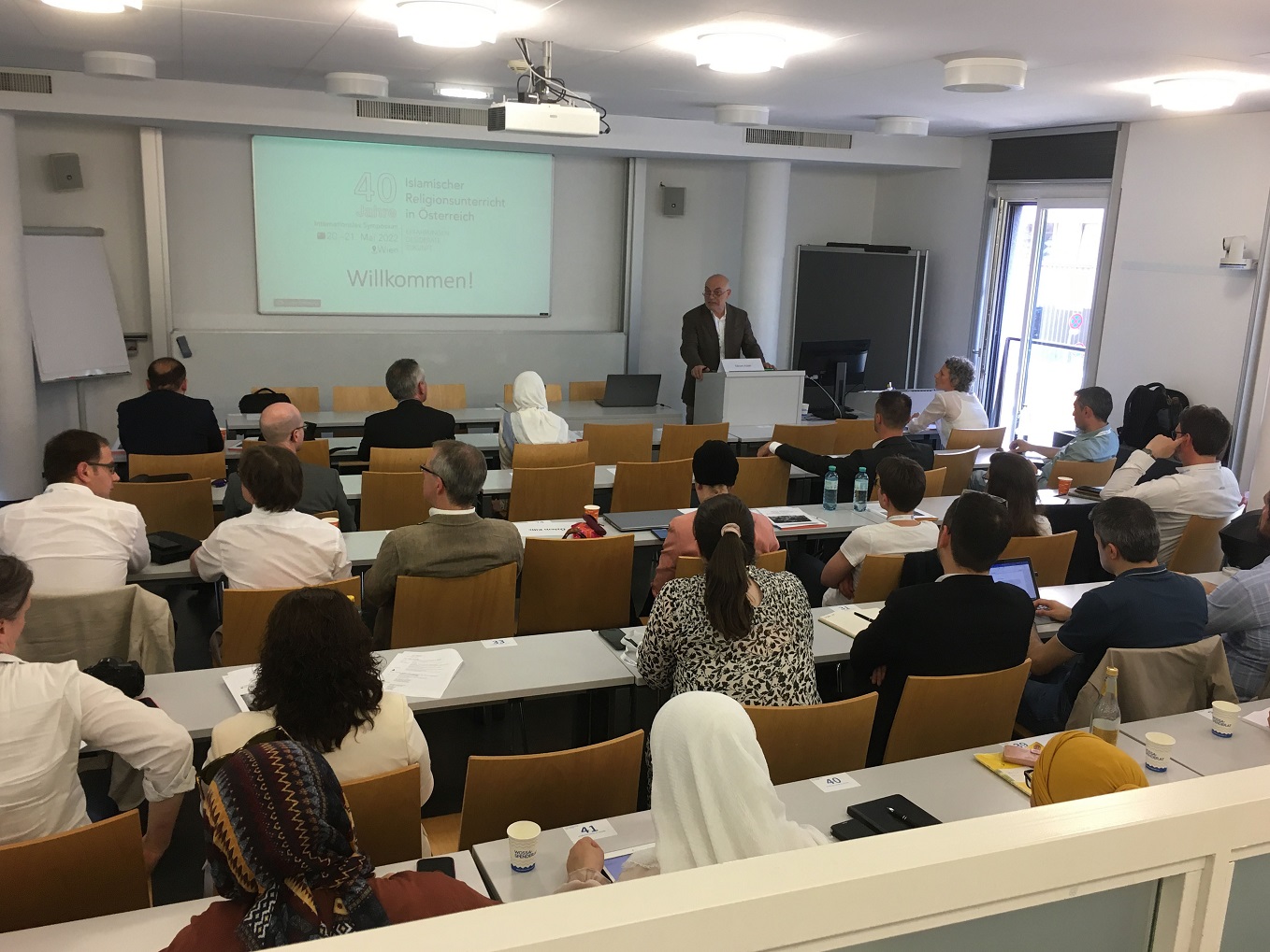 Bild: Foto von Prof. Ednan Aslan am Rednerpult, daneben eine Powerpoint-Präsentation; davor die zuhörenden Teilnehmenden im Hörsaal